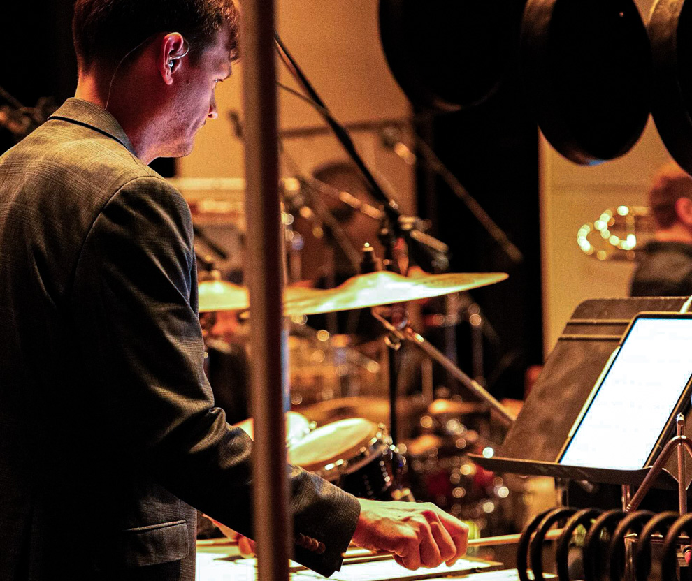 A musician in a suit plays percussion instruments on stage, reading sheet music from a stand, with drums and microphones visible in the background.