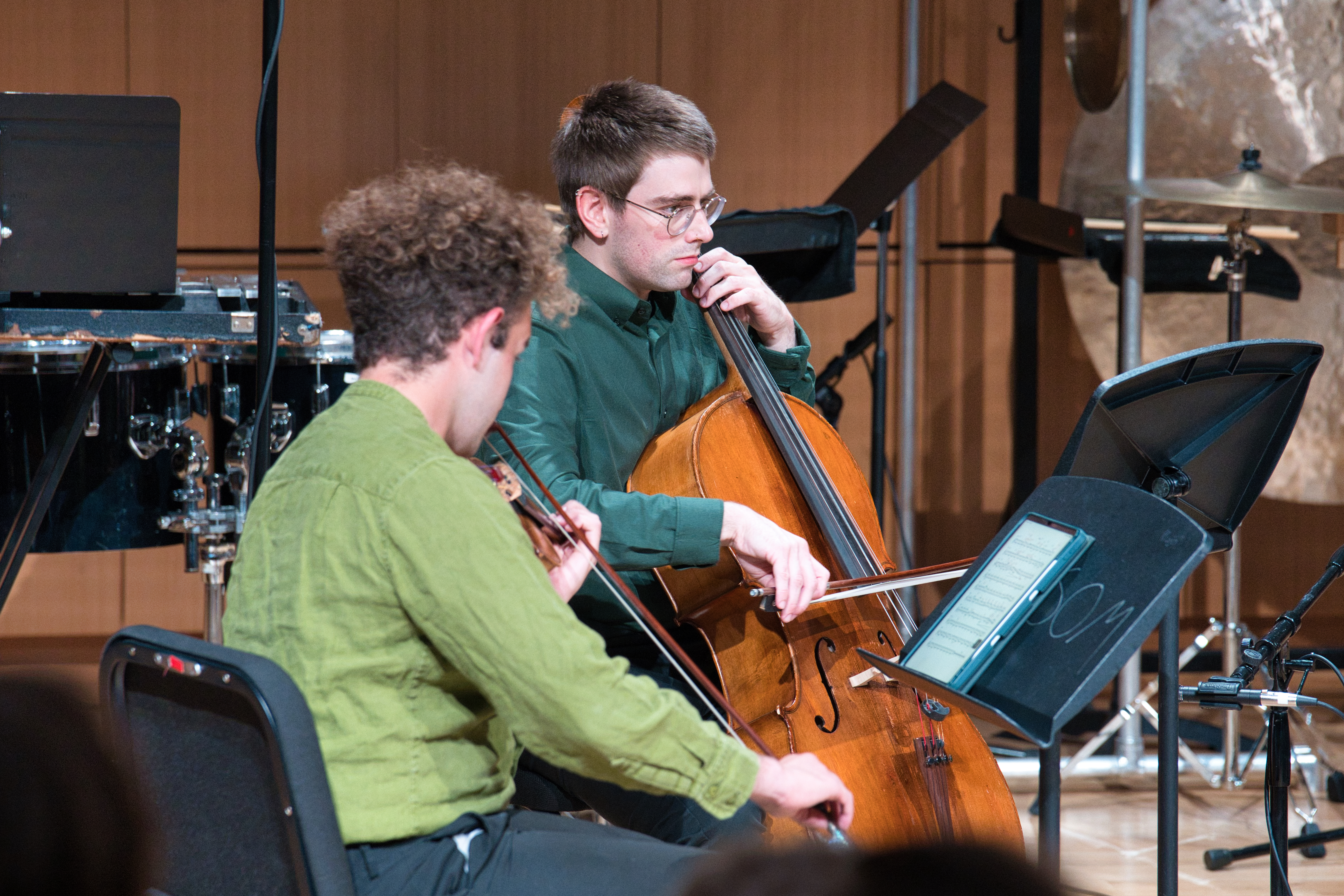 Two musicians perform on stage with a violin and cello. Tablet with music is visible on a stand in the foreground.
