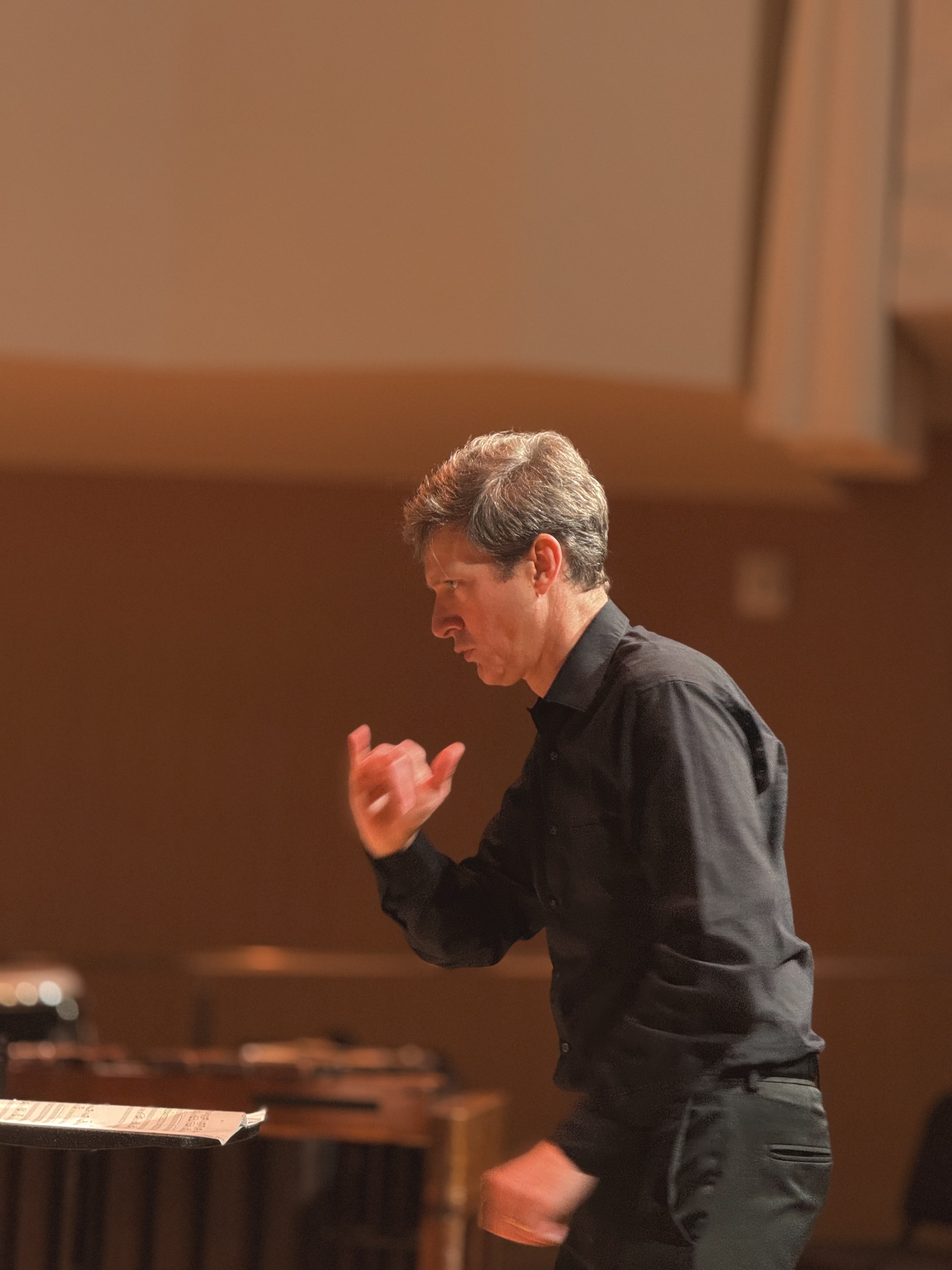 A conductor in a black shirt passionately directs musicians, gesturing with his hands. The warm lighting and blurred background suggest a concert setting.