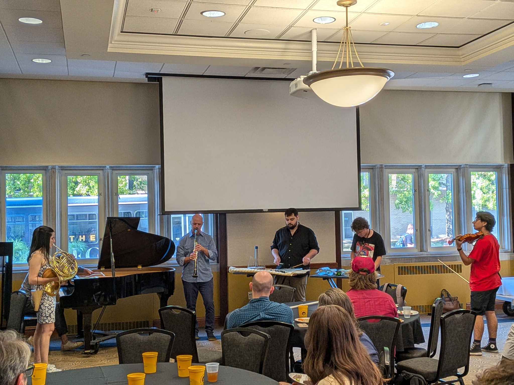 A group of musicians performs in a room with a piano and various instruments, watched by seated attendees. The atmosphere is casual and engaged.