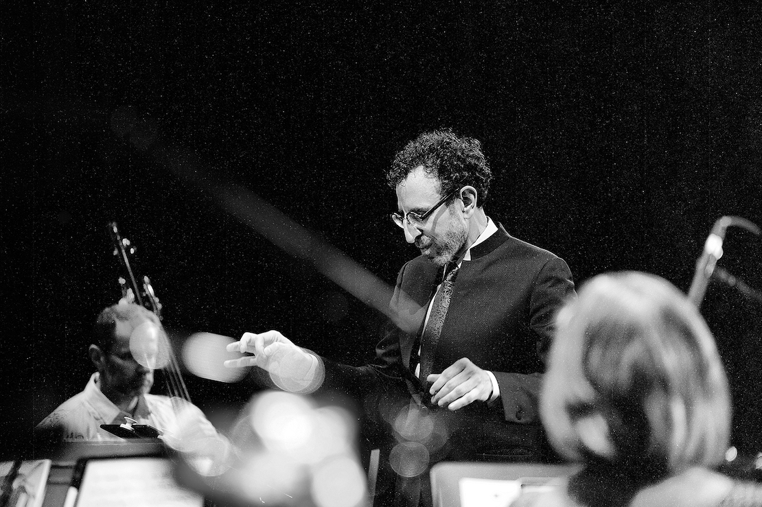 A conductor in a suit passionately leads a chamber orchestra, hands poised mid-motion. Musicians and instruments, blurred, frame the background. Black and white.