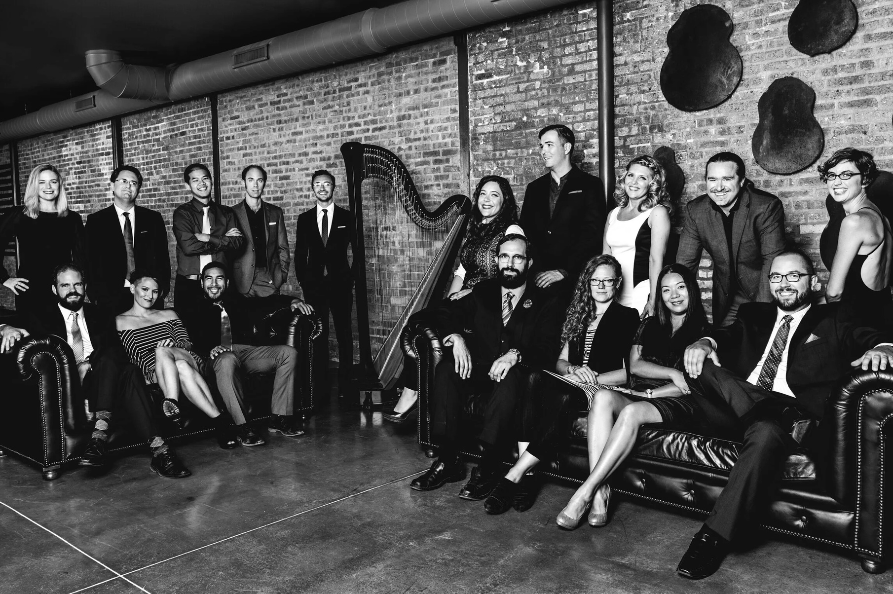 Black-and-white group portrait of a formally dressed ensemble in a brick-walled loft, gathered around a harp and leather couches, smiling warmly.