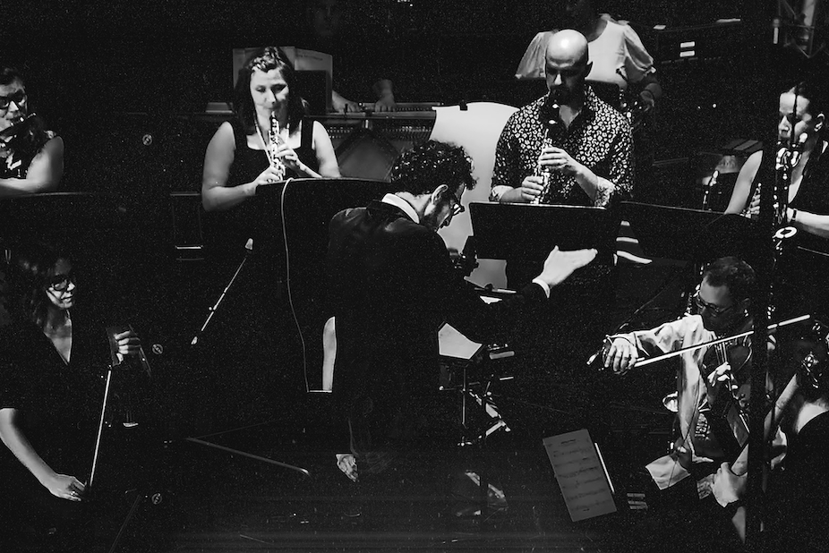 A black-and-white image of an orchestra performing. A conductor stands in the center, surrounded by musicians playing violins, clarinets, and cellos, creating an intense and dramatic atmosphere.