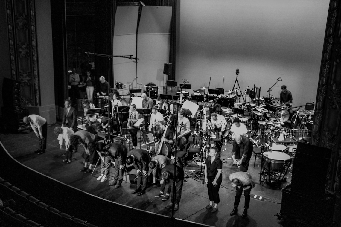 Black and white image of a crowded stage with musicians bowing towards the audience. Instruments and equipment are scattered around.