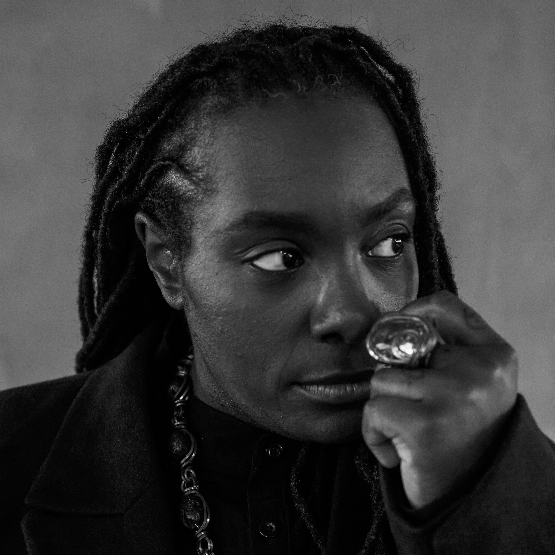 A person with long braided hair gazes thoughtfully to the side. Wearing a dark coat and large ring, the expression is introspective and calm. Black and white.