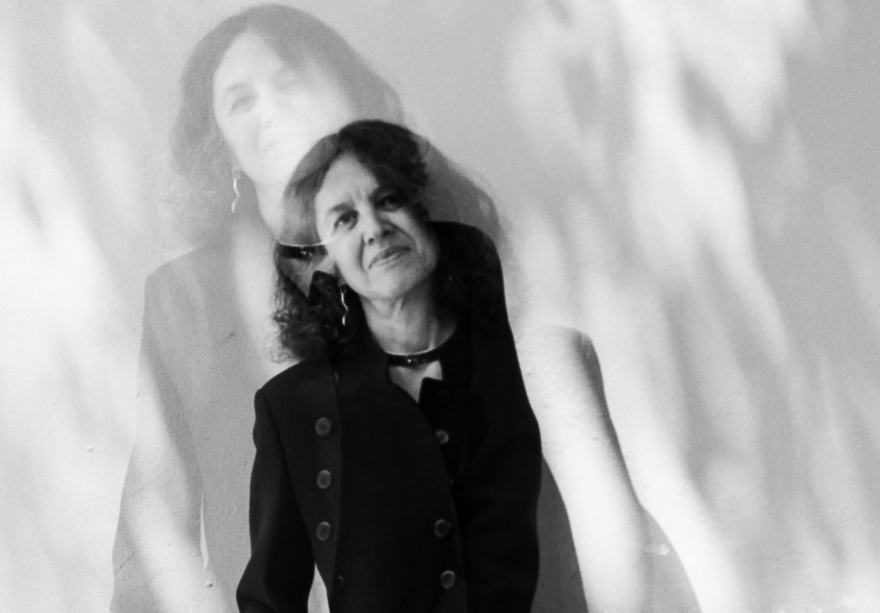 A woman with long hair leans casually against a textured wall, wearing a dark suit. Her image in a slightly different pose is subtly superimposed.
