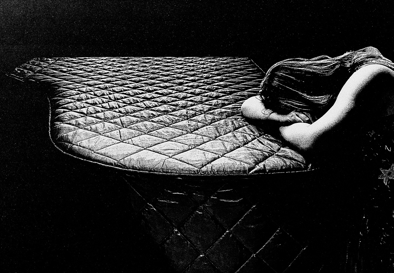 A person with long hair leans forward, arms crossed over the quilted, textured surface of a piano cover, in a dark, shadowy setting, conveying a feeling of solitude and emotional intensity.