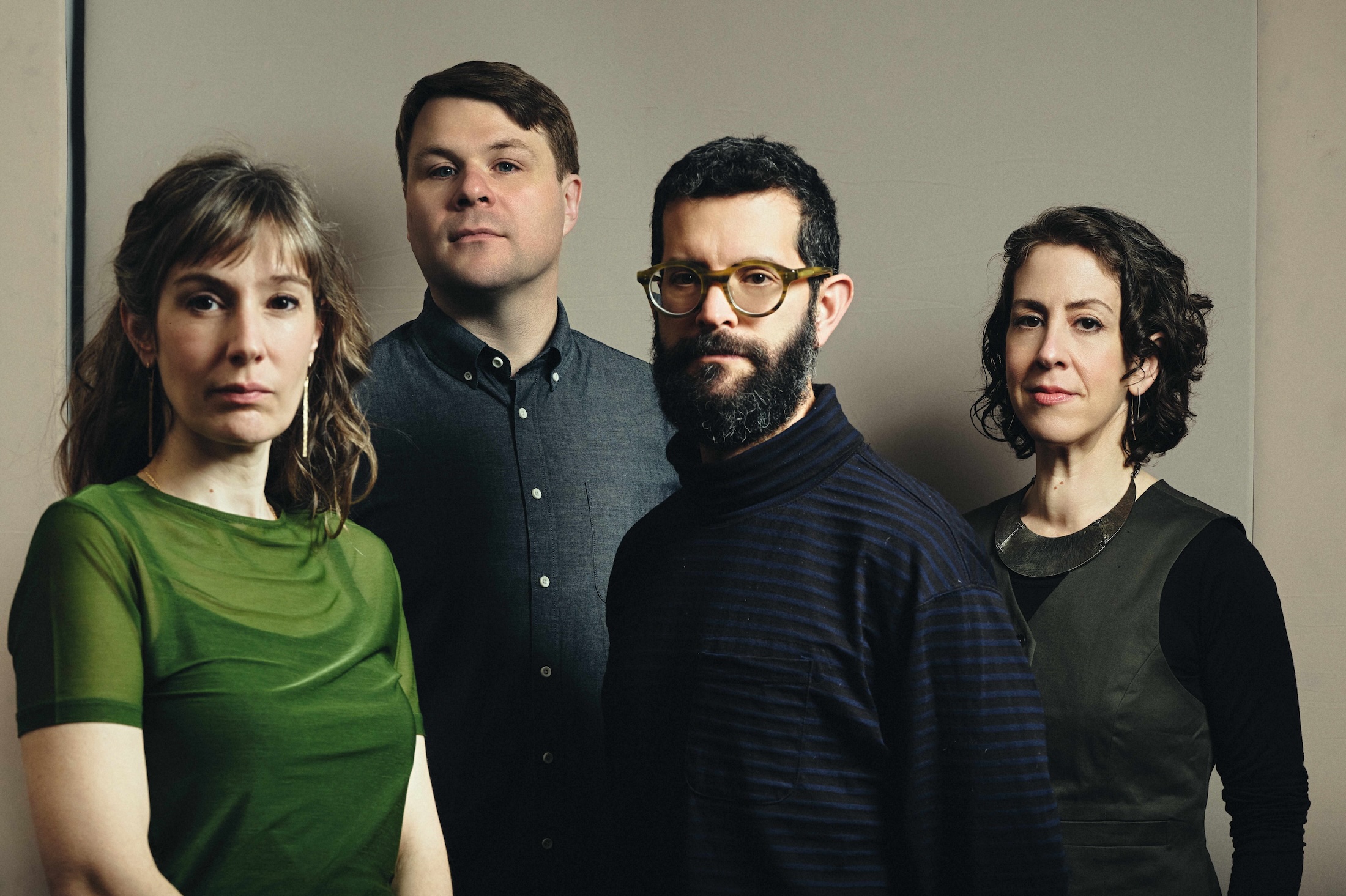 A group of four adults stands closely together, each with a neutral expression. They are dressed in casual, muted clothing against a plain background.