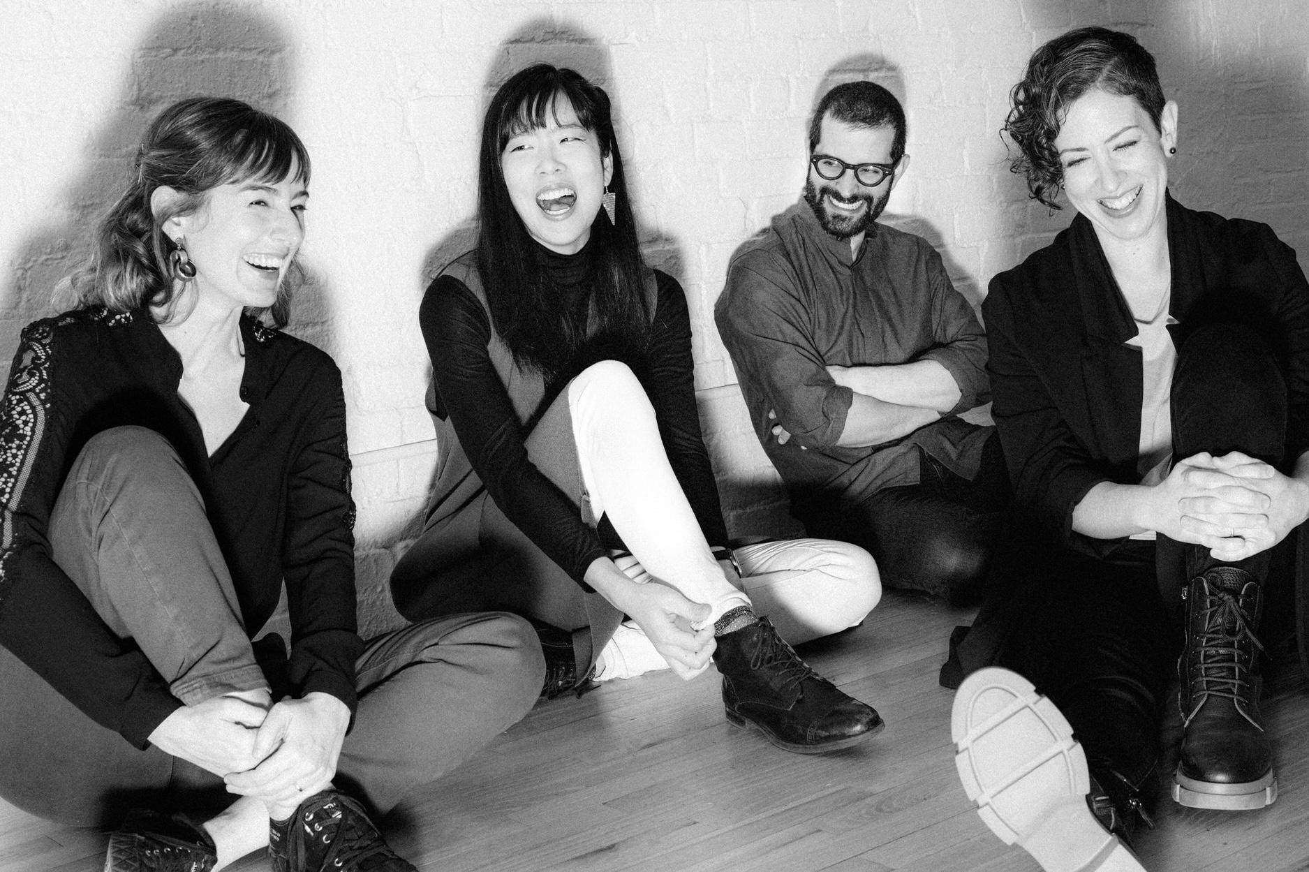 A black and white image of four people sitting on a wooden floor, laughing and relaxed. They lean against a white brick wall, exuding joy and camaraderie.