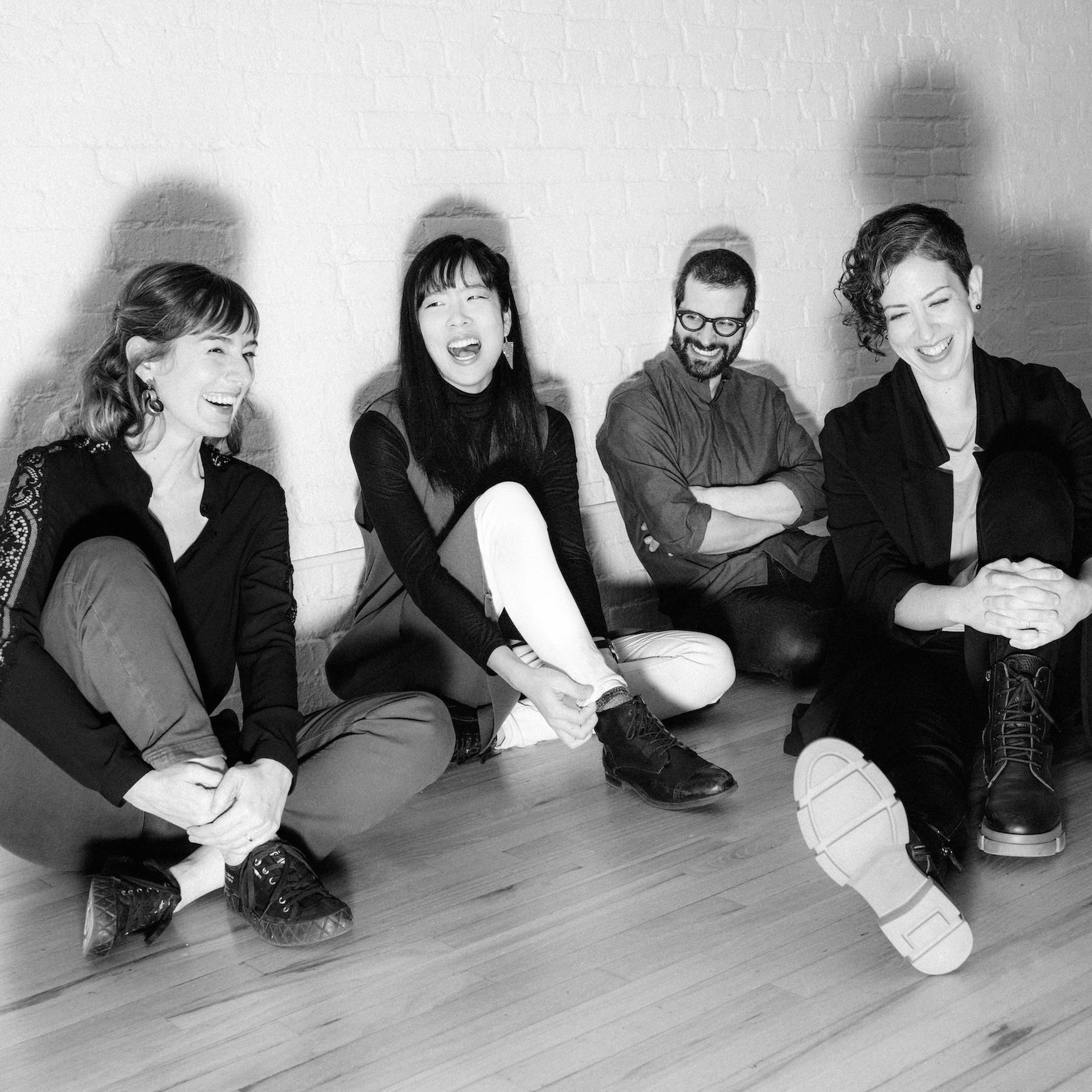 A black and white image of four people sitting on a wooden floor, laughing and relaxed. They lean against a white brick wall, exuding joy and camaraderie.