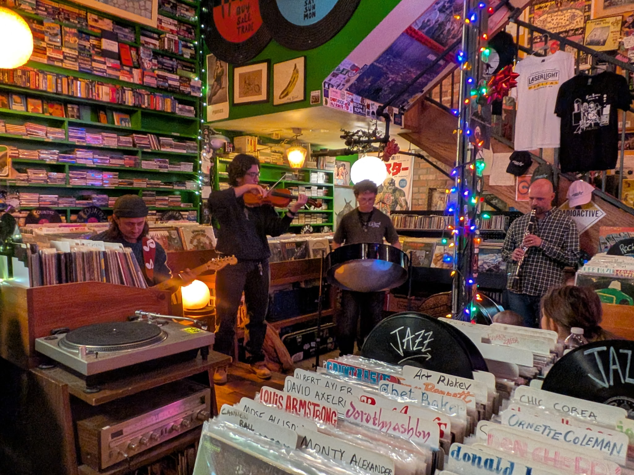 A lively scene in a cozy record store, with four musicians playing guitar, violin, steel drum, and clarinet. Vinyl records and CDs line the walls, illuminated by warm lights and colorful string lights. The atmosphere is vibrant and slightly nostalgic.