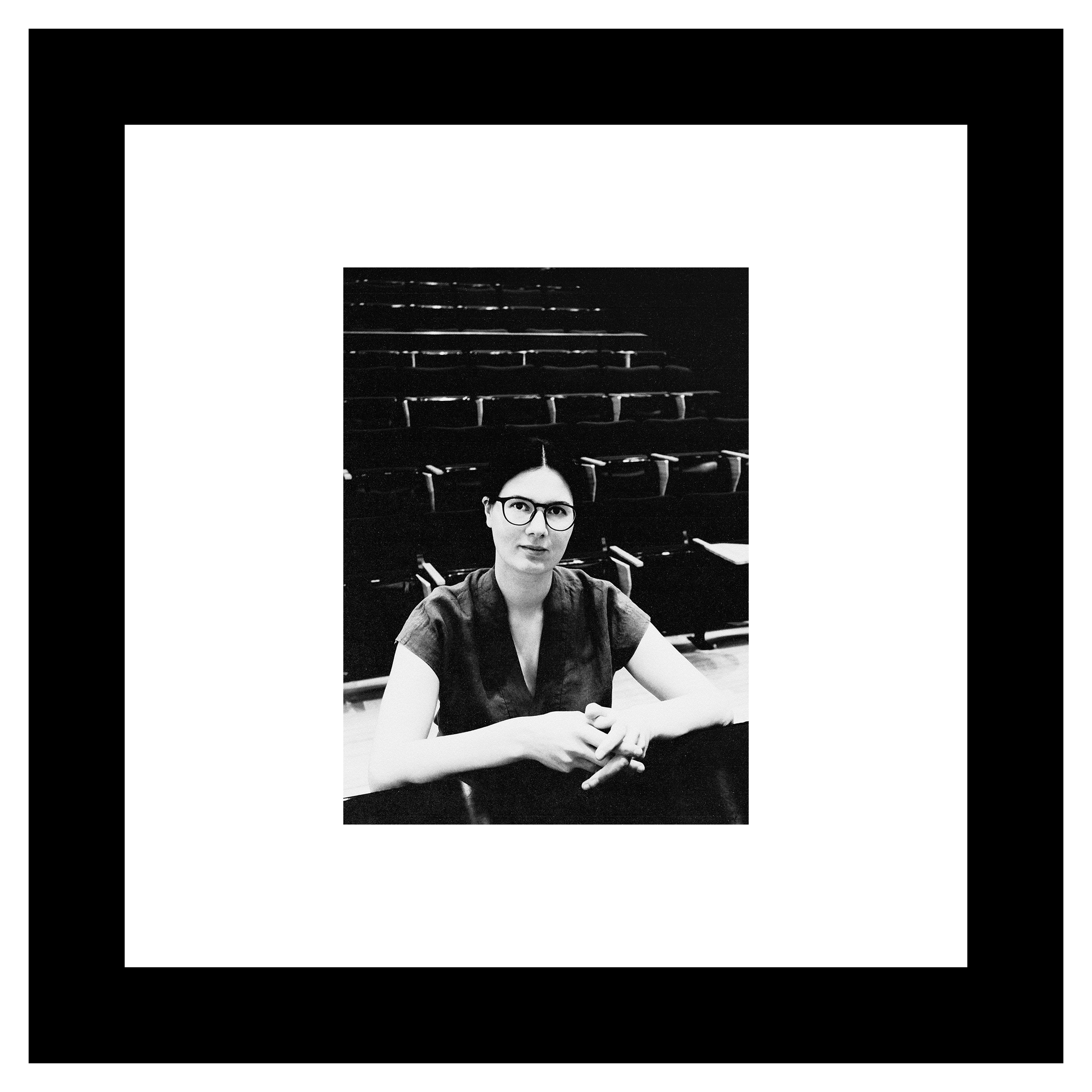 Inga Chinilina photo. Black and white photo of a person with glasses, seated in a lecture hall. They appear thoughtful, surrounded by empty chairs.