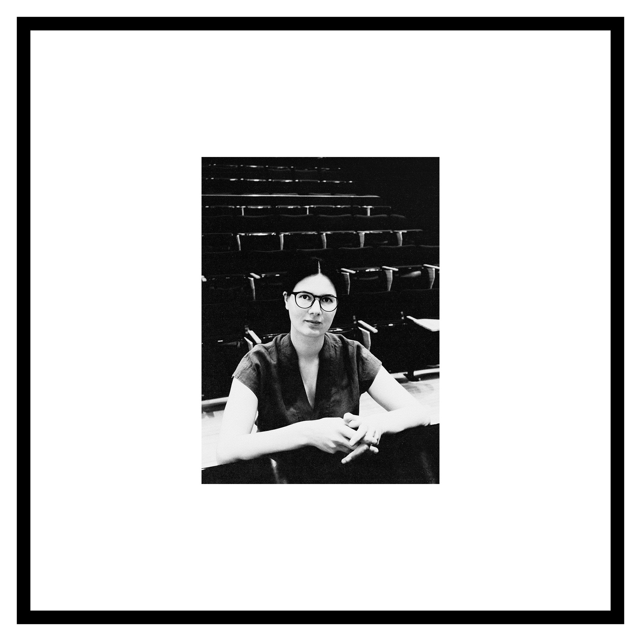 Inga Chinilina photo. Black and white photo of a person with glasses, seated in a lecture hall. They appear thoughtful, surrounded by empty chairs.
