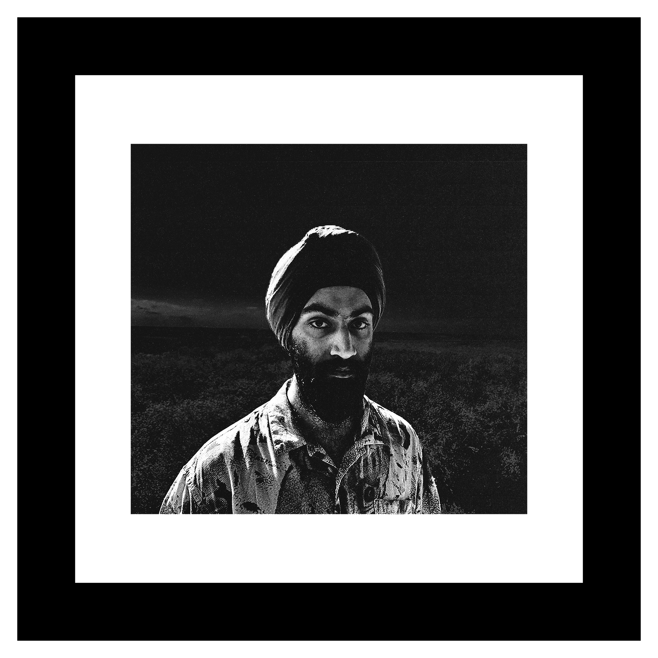 Arjan Singh Dogra portrait. Black and white portrait of a man wearing a turban and patterned shirt, standing against a dark, blurred landscape. The expression appears serious.