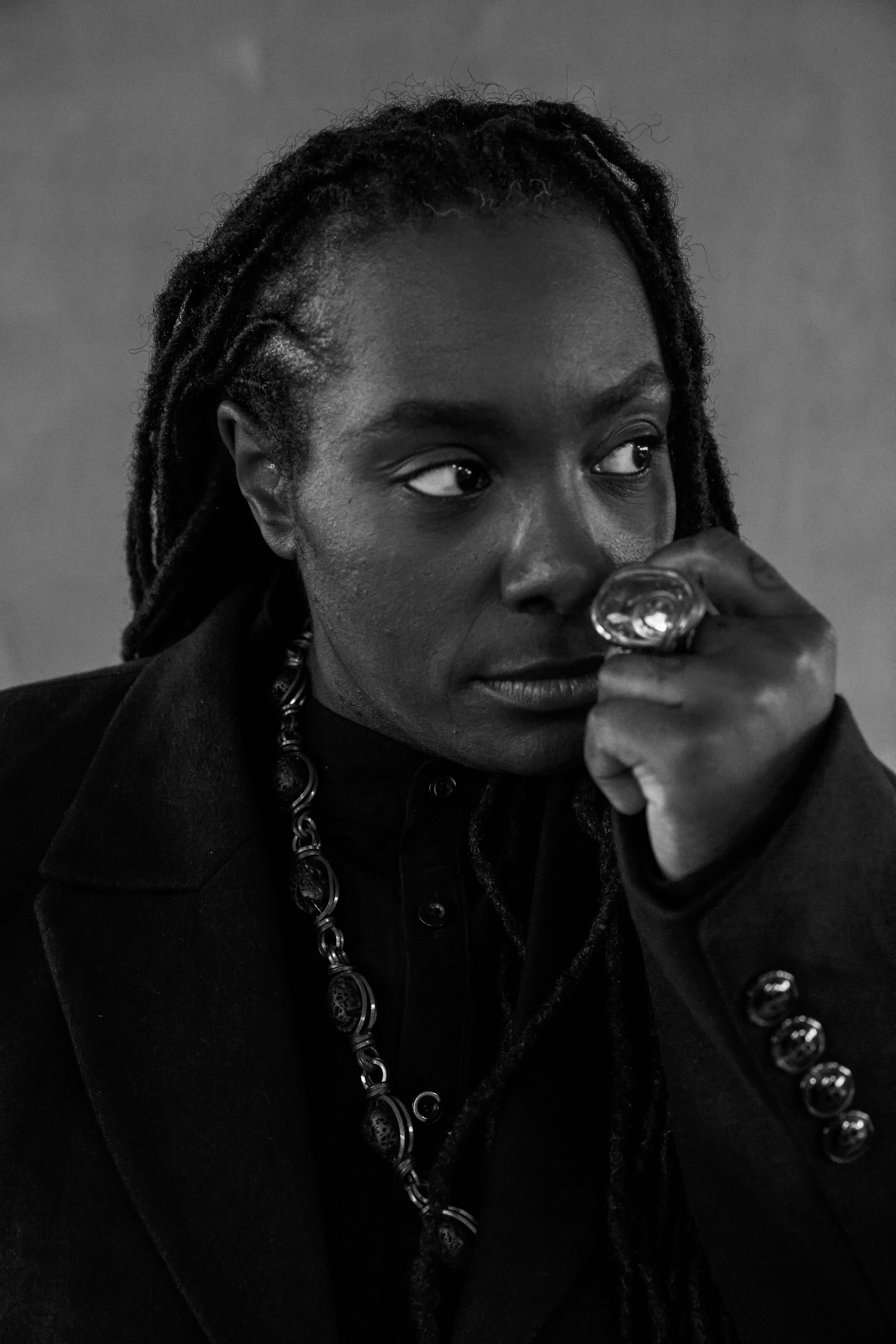 A black and white close-up of a woman with long locs and thoughtful expression. She wears a dark coat, a thick necklace, and a large, ornate ring, conveying introspection.