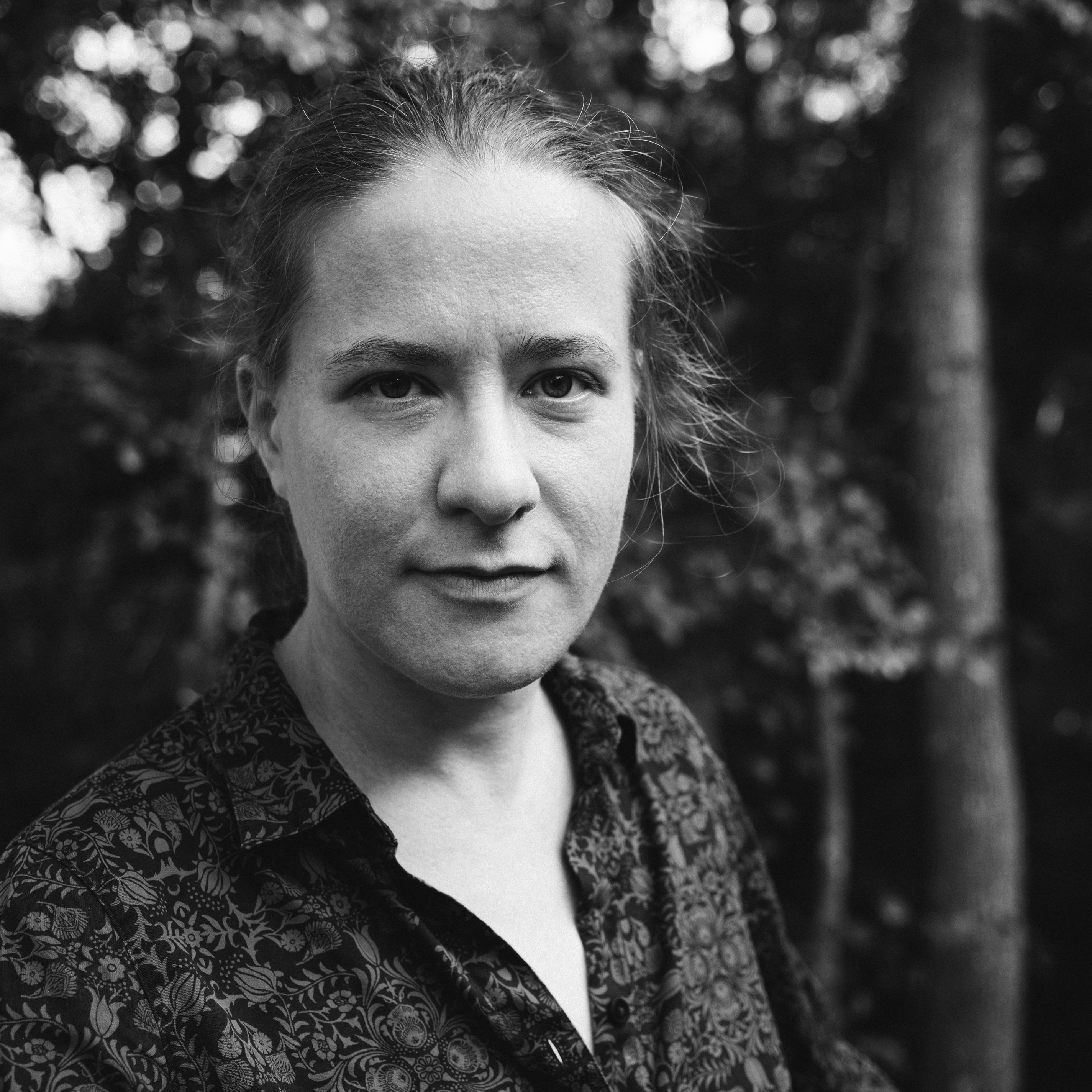 Black and white portrait of a person with a serious expression, wearing a patterned shirt, standing in a wooded area with soft, blurred trees behind.