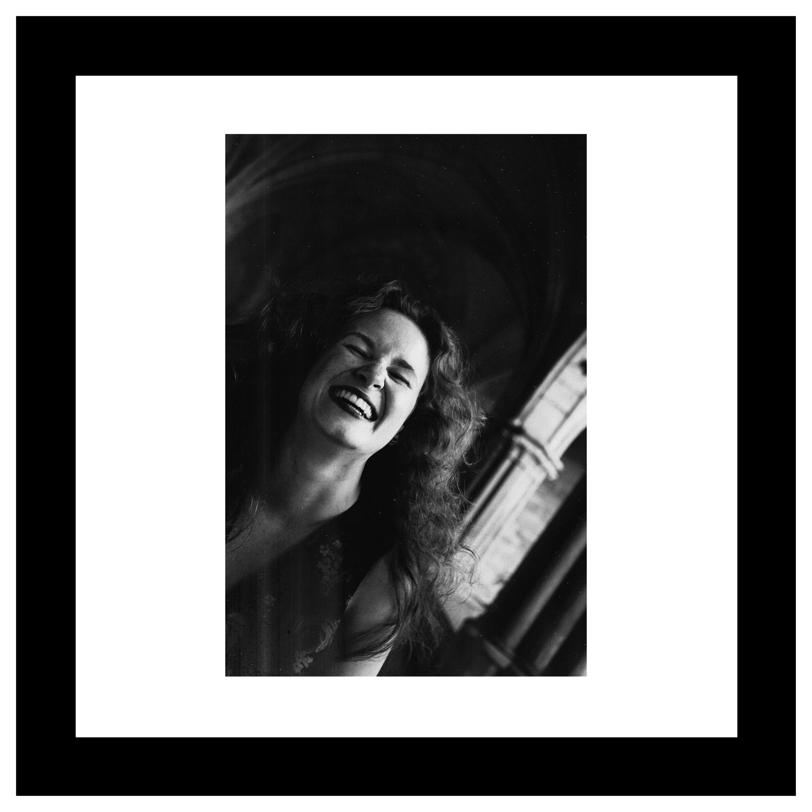 Ihlara McIndoe portrat. Black and white photo of a woman with curly hair laughing openly. Background features a softly lit archway, conveying joy and spontaneity.