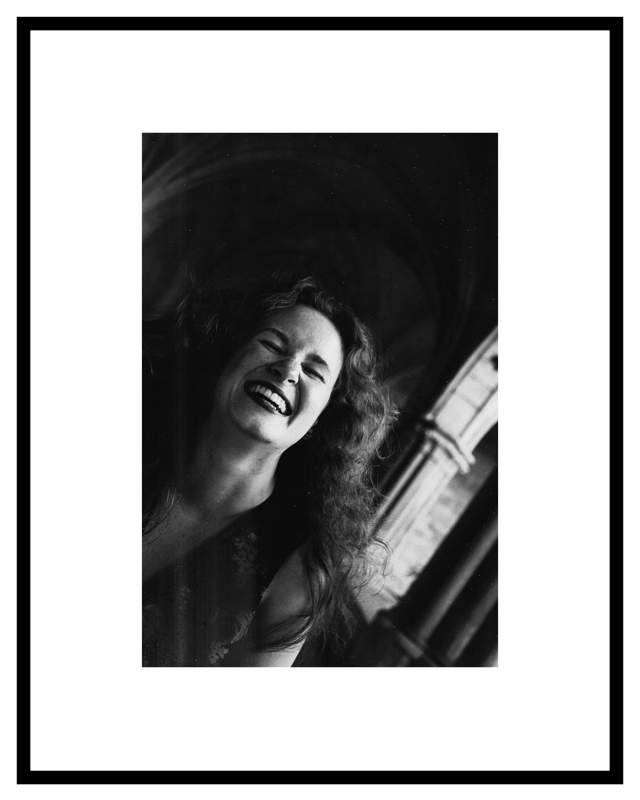 Ihlara McIndoe portrat. Black and white photo of a woman with curly hair laughing openly. Background features a softly lit archway, conveying joy and spontaneity.