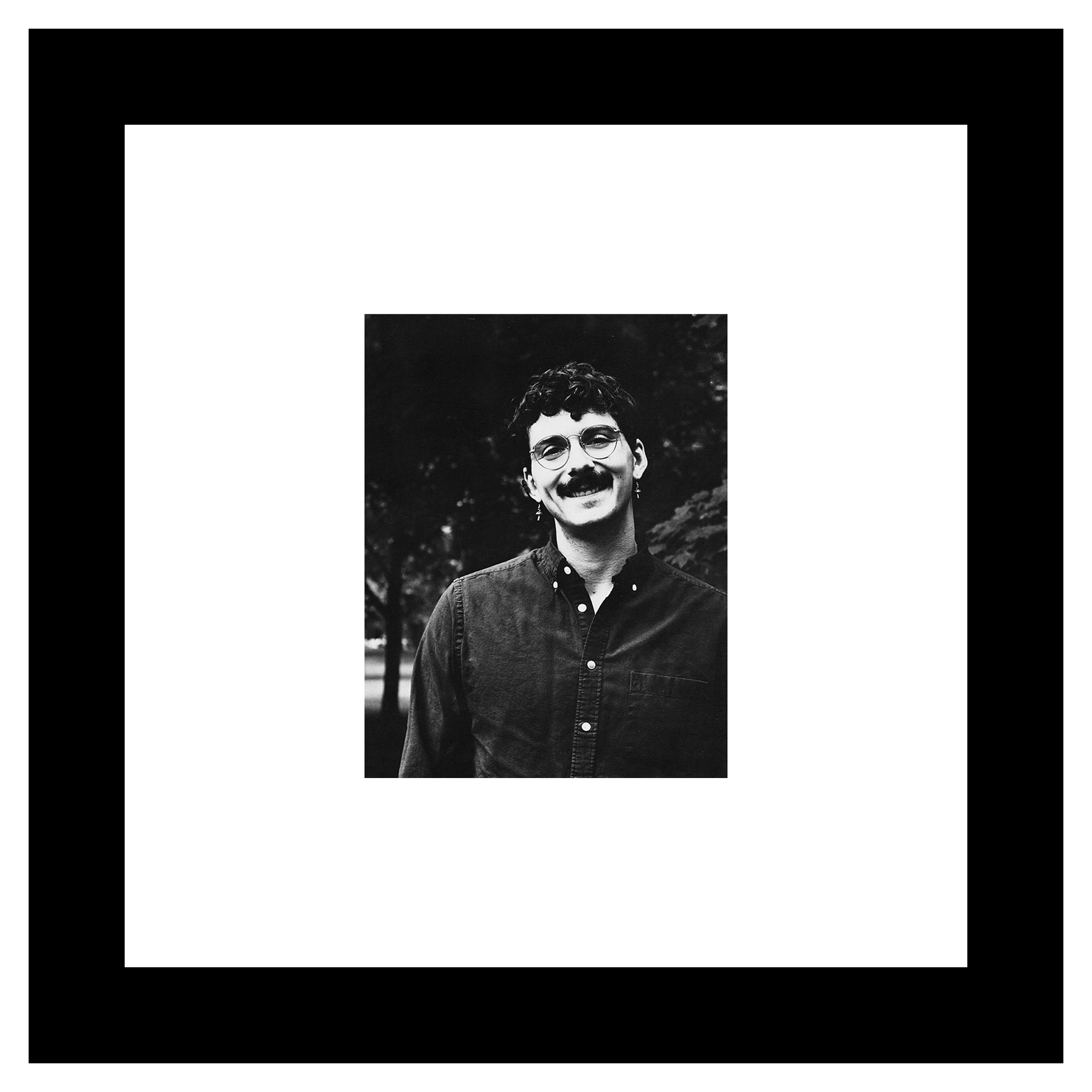 Paul Novak portrait. Black and white photo of a person with curly hair, glasses, and a thick mustache, smiling warmly in a dark, leafy outdoor setting.