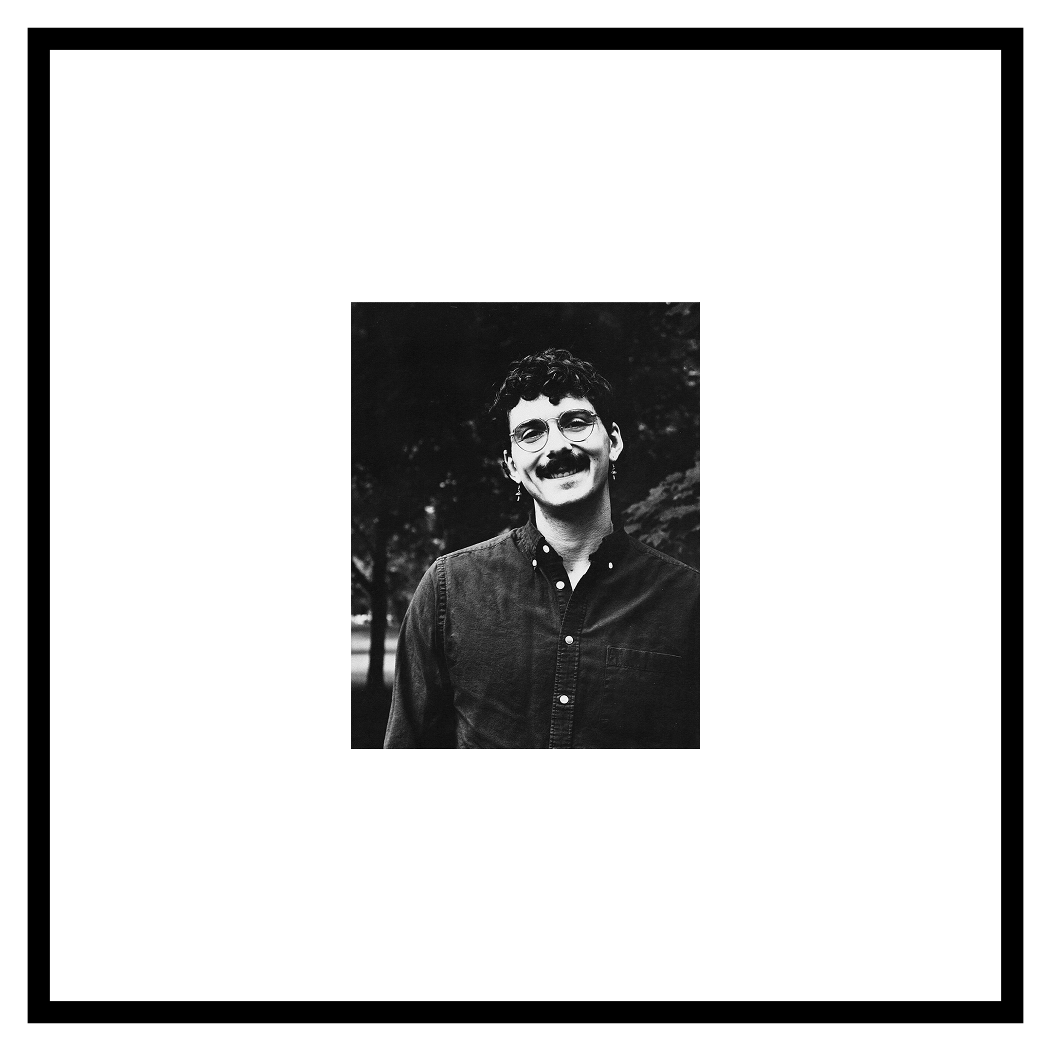 Paul Novak portrait. Black and white photo of a person with curly hair, glasses, and a thick mustache, smiling warmly in a dark, leafy outdoor setting.