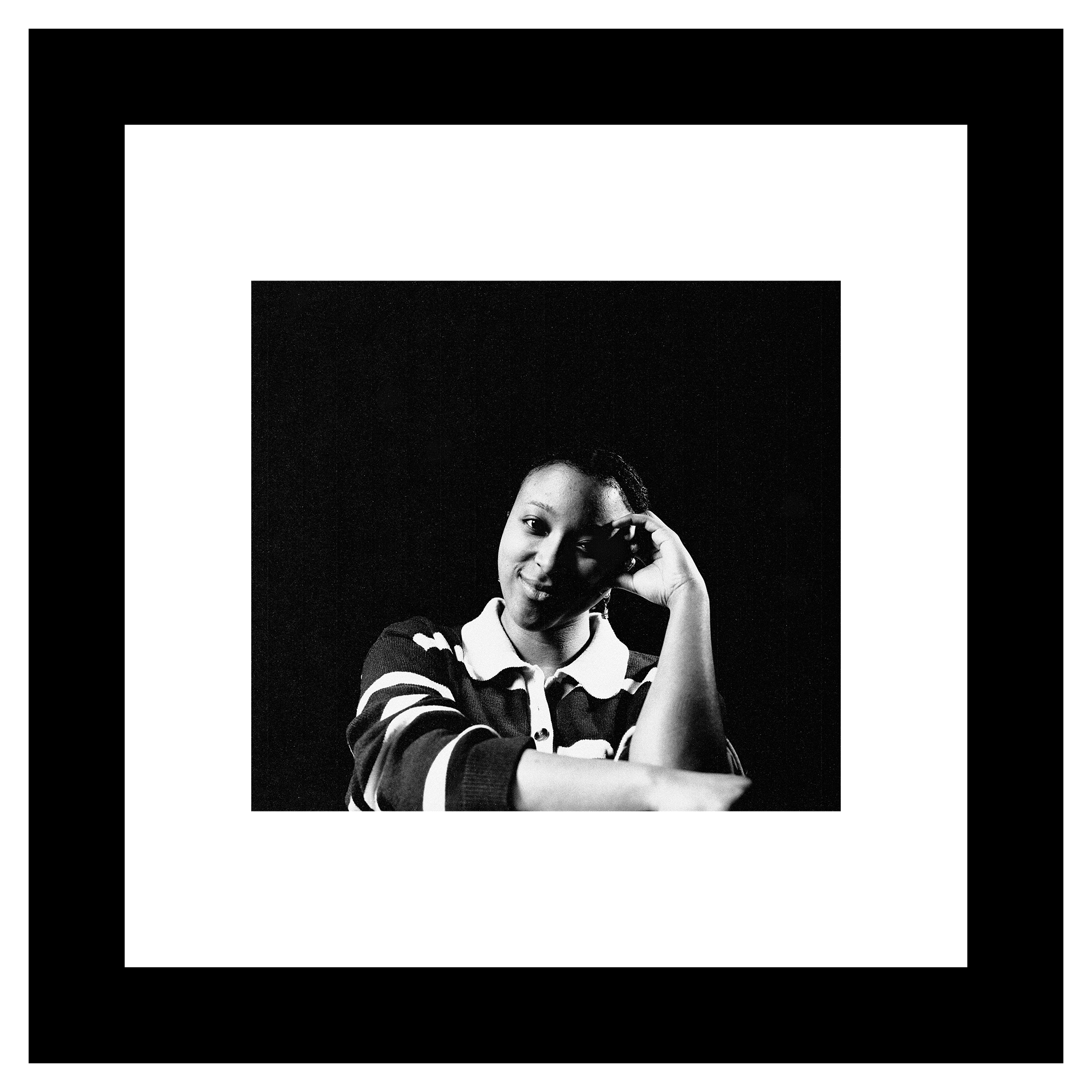 Danae Venson portrait. A woman in a striped shirt sits against a dark background, resting her temple on her hand with a thoughtful and serene expression. Black and white tone.