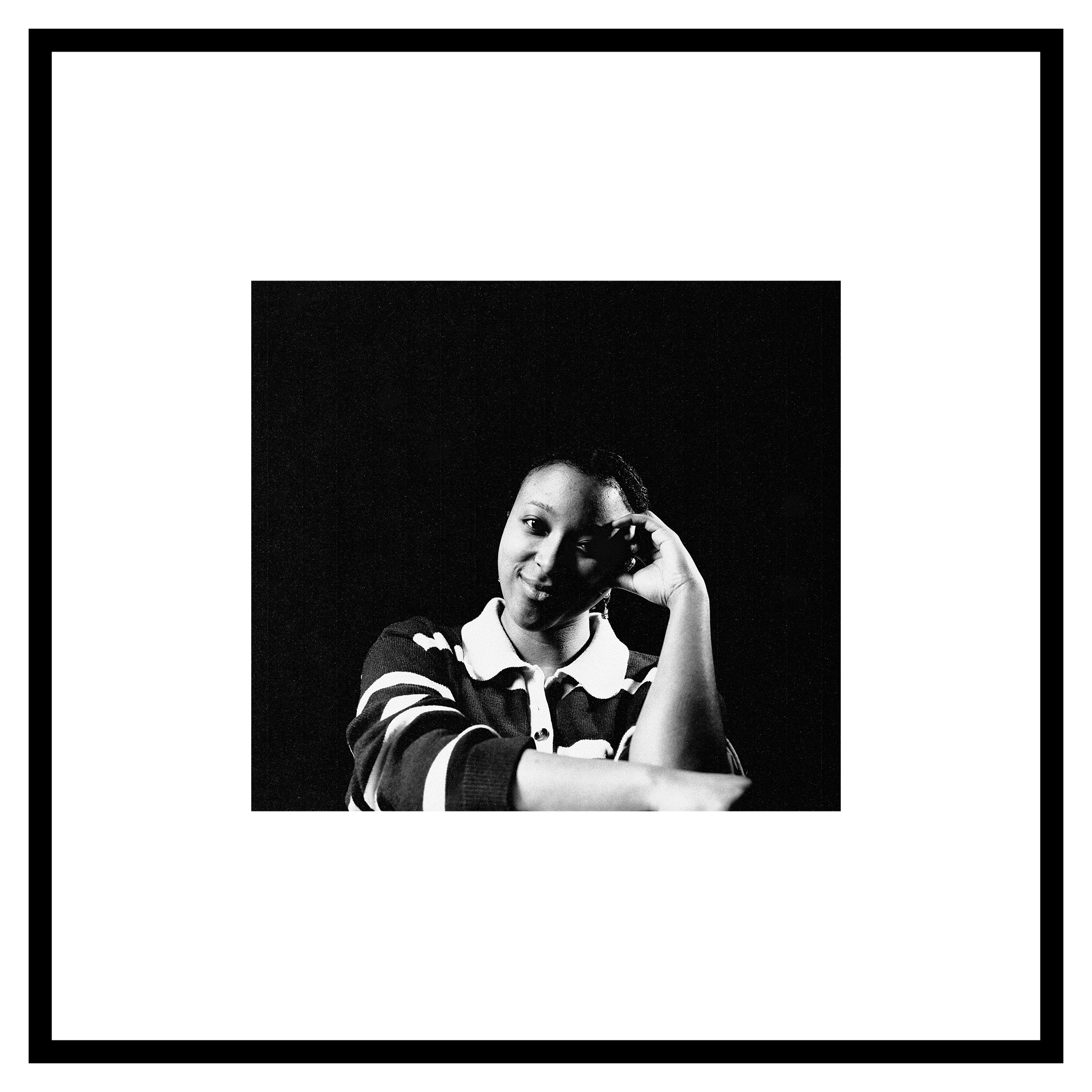 Danae Venson portrait. A woman in a striped shirt sits against a dark background, resting her temple on her hand with a thoughtful and serene expression. Black and white tone.