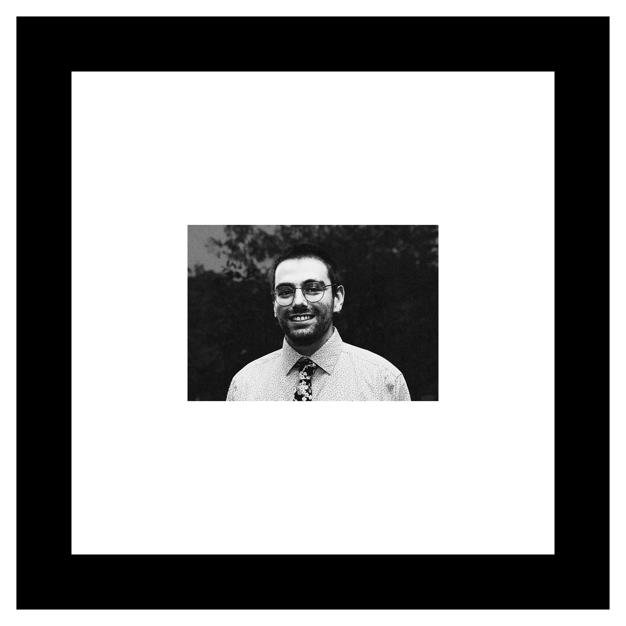 Dean Wibe portrait. A man with a broad smile and glasses, a dark patterned tie on a patterned shirt, and a 5 o'clock shadow stands in front of a blurred background of trees and sky. Black and white.