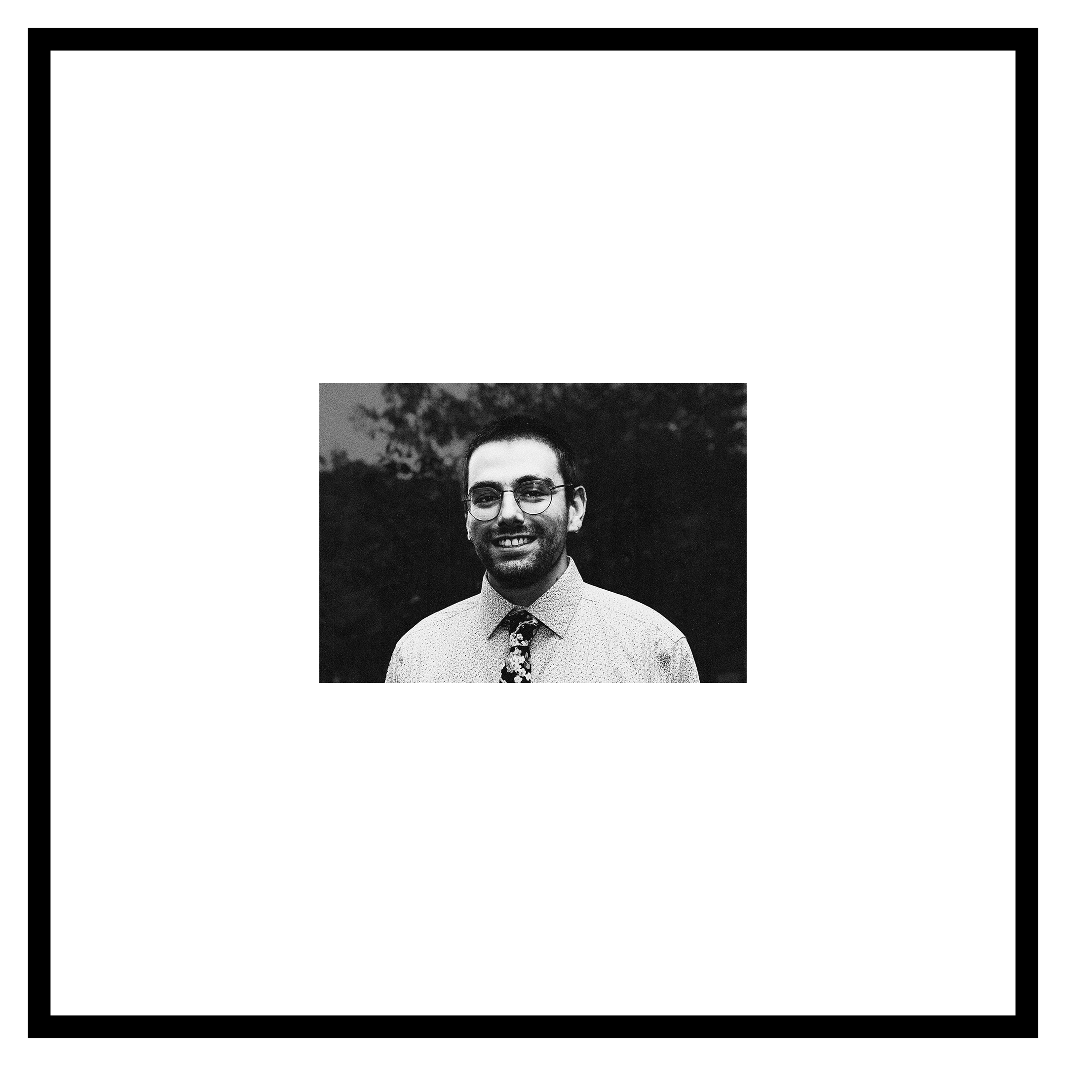 Dean Wibe portrait. A man with a broad smile and glasses, a dark patterned tie on a patterned shirt, and a 5 o'clock shadow stands in front of a blurred background of trees and sky. Black and white.