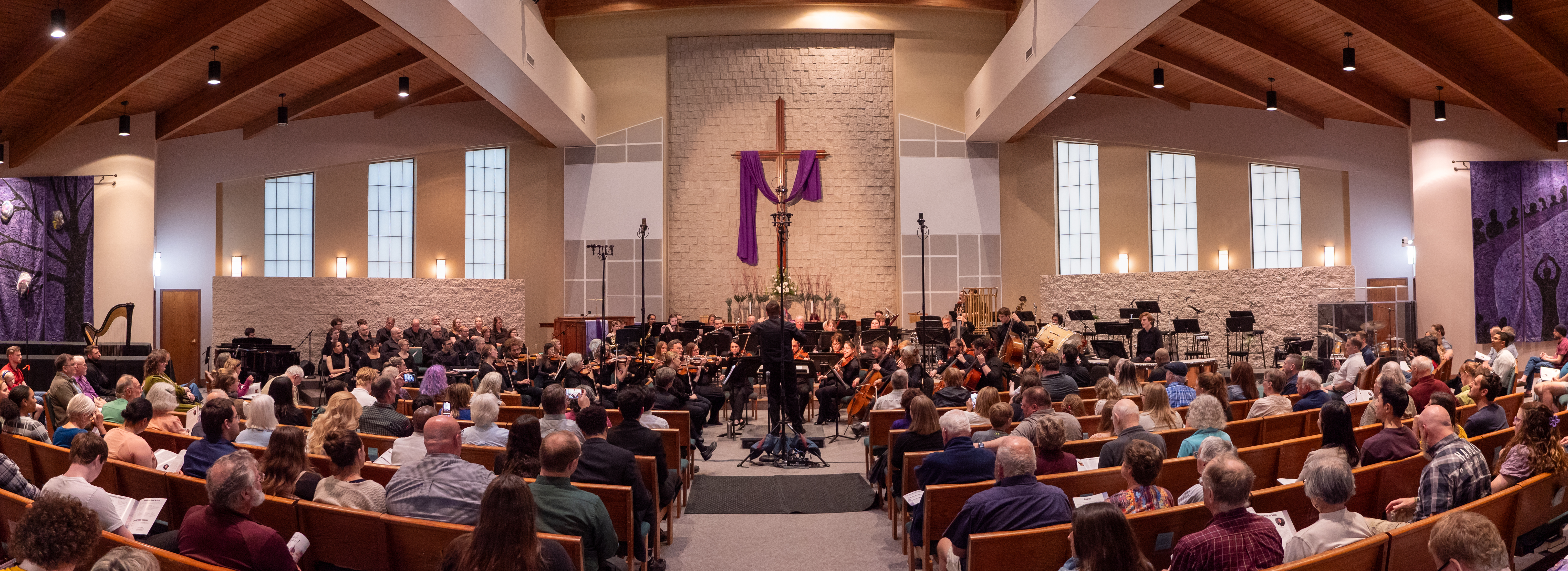 A full orchestra performs in a church. The space has wooden beams and purple drapery. Lighting is soft but not dim. A cross is displayed, and a large audience is seated, creating a contemplative atmosphere.