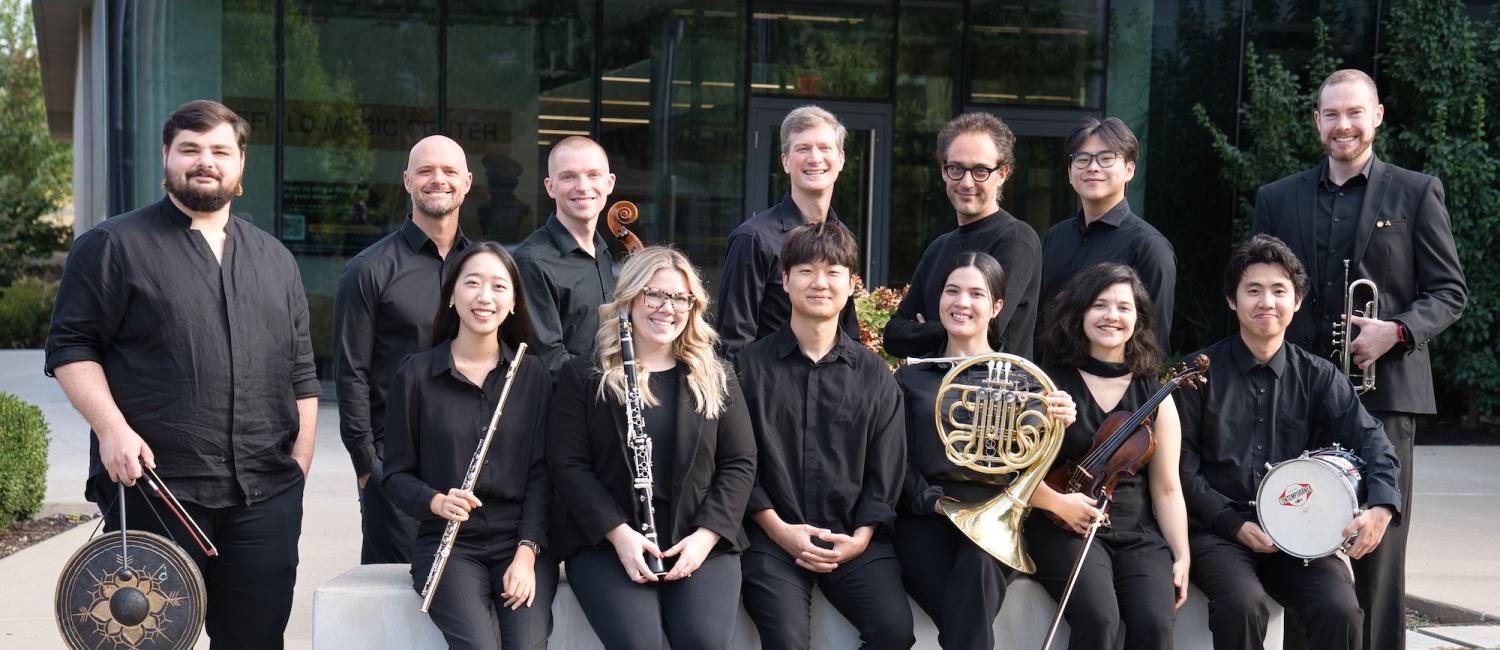 A group of twelve musicians poses outdoors, wearing black attire. They hold various instruments, including a gong, flute, oboe, horn, and violin, exuding a formal and united tone. This is the 2025-2026 New Music Ensemble.