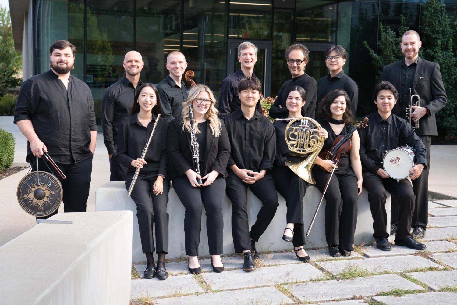A group of 12 musicians, dressed in black, pose outside a modern building, the Sinquefield Music Center on Mizzou's campus. They hold various instruments including a flute, French horn, and trumpet, smiling warmly. This is the 2025-2026 New Music Ensemble with director Stefan Freund.